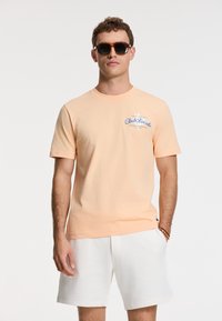 Lys orange bomulds T-shirt med rund hals, korte ærmer og blå grafisk tekst på brystet. Sammen med hvide shorts og solbriller.