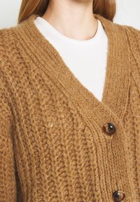 Brauner gestrickter Cardigan mit strukturiertem Muster, tiefem V-Ausschnitt, zwei großen Knöpfen und einer weichen, voluminösen Verarbeitung.