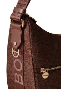 Braune synthetische Schultertasche mit strukturiertem Finish, goldfarbener Hardware, einem gemusterten Trageriemen und einer Reißverschlusstasche an der Seite.