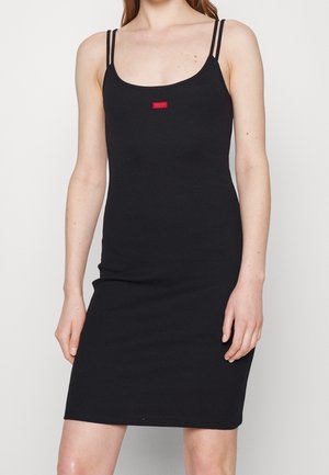 Woman wearing a fitted black sleeveless dress with thin double straps and a small red logo on the chest.
