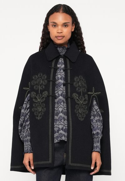 Μαύρη μάλλινη κάπα με λουλουδάτο κέντημα, κούμπωμα με κουμπιά, και μυτερό γιακά. Διαθέτει μοτίβο στην εσωτερική επένδυση και υφασμάτινα μανίκια.