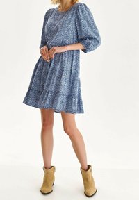 Blaues Blumenkleid mit puffartigen Ärmeln und gestuftem Saum. Hergestellt aus leichtem Material, hat es ein strukturiertes Muster und wird mit braunen Knöchelschuhen kombiniert.