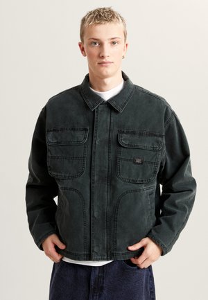 Ciemnozielona kurtka jeansowa z kołnierzykiem, dwoma kieszeniami na piersi oraz subtelnymi przeszyciami, noszona na białej koszulce.