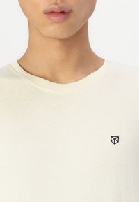 Kremasta majica od mekanog pamuka, s okruglim izrezom i malim crnim geometrijskim logotipom s lijeve strane prsiju.
