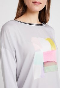 Hellgraues Langarmoberteil mit einem schwarz gestreiften Ausschnitt, das ein buntes abstraktes Design in Rosa, Gelb, Grün und Blau auf der Vorderseite aufweist.