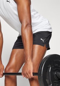 Svarta atletiska shorts med vitt Puma-logotyp, kombinerat med en vit linne. Visar armar som greppar en svart viktstång. Fokus på fitnesskontext.