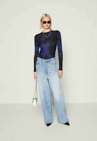 Γυναίκα που φοράει μαύρη και μπλε πετσετέ μπλούζα με μακριά μανίκια, ανοιχτό μπλε φαρδύ παντελόνι τζιν, μαύρες μυτερές γόβες, γυαλιά ηλίου, αλυσίδα στο λαιμό, κρατώντας λευκή τσάντα.