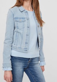 Veste en denim bleu clair avec poches avant, boutons en métal argenté et design ajusté. Portée sur un haut assorti en bleu clair avec des poignets côtelés.