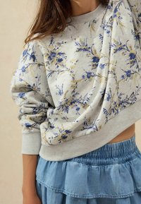 Sweat-shirt gris à motif floral avec accents bleu et or. Doté d'une coupe décontractée, d'un col rond et de poignets élastiqués, associé à une jupe bleue superposée.