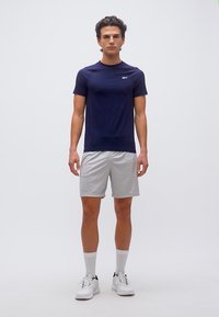 Νεαρός άνδρας που στέκεται φορώντας μπλουζάκι αθλητικό ναυτικού μπλε χρώματος, ανοιχτό γκρι σορτς, λευκές κάλτσες και λευκά αθλητικά παπούτσια σε απλό λευκό φόντο.