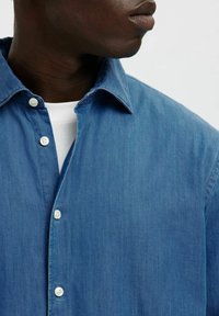 Ljusblå denimskjorta med klassisk krage, knappstängning fram med vita knappar och en slät yta. Synlig sömnad i kanterna.