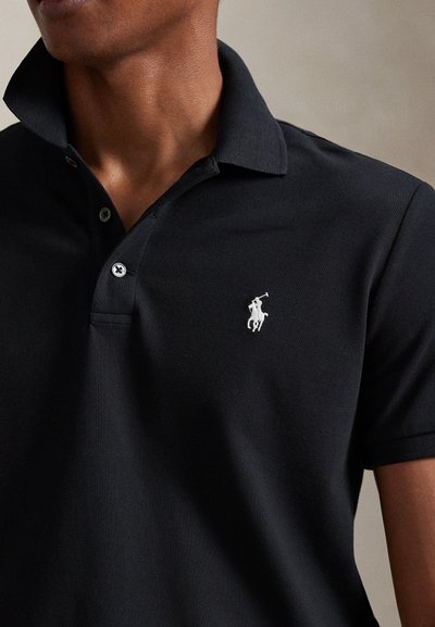 Κοντινό πλάνο ενός ατόμου που φοράει μαύρο πόλο με λευκό κεντημένο λογότυπο παίκτη πόλο στο αριστερό στήθος.