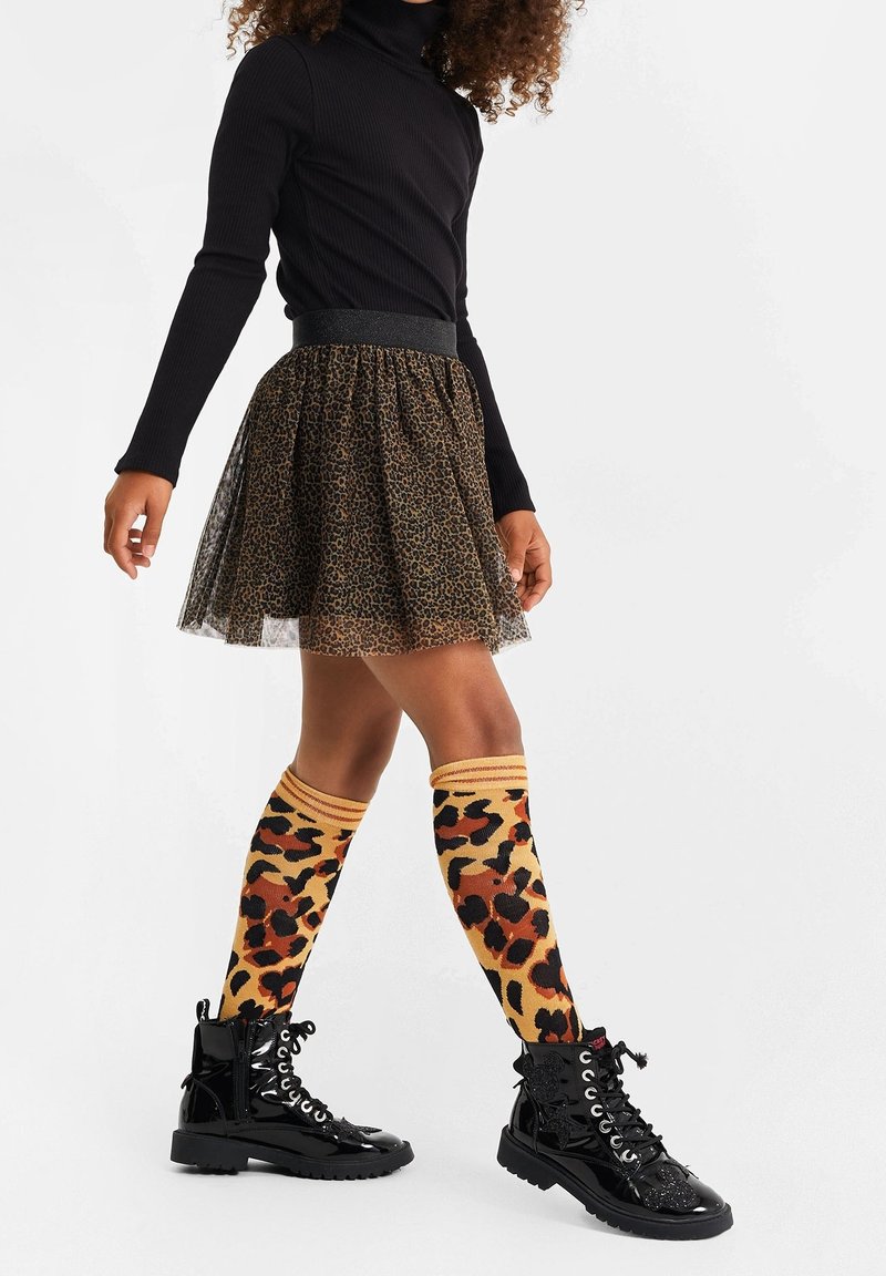 Personne portant un haut noir à manches longues, une jupe imprimé léopard, des chaussettes montantes assorties à imprimé léopard et des bottes noires vernies à lacets.