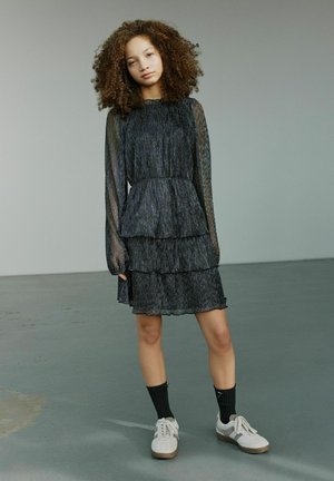 Schwarzes, gestuftes Kleid aus transparentem, plissiertem Stoff, mit langen Ärmeln und einem gerafften Kragen, kombiniert mit weißen Sneakern und schwarzen Socken.