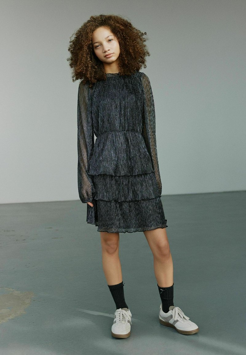 Čierne, vrstvené šaty z priehľadnej plisovanej látky, s dlhými rukávmi a volánikovým výstrihom, v kombinácii s bielymi teniskami a čiernymi ponožkami.