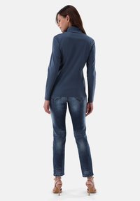 Langärmliges, dunkelgraues Oberteil mit hohem Kragen, kombiniert mit verwaschenen blauen Jeans; das Model trägt hellrosa Absätze. Glatter Stoff, schlanke Passform.