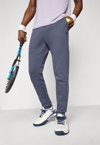 Person trägt graue Jogginghose und weiße Tennisschuhe, hält in einer Hand einen Tennisschläger und in der anderen einen Tennisball.