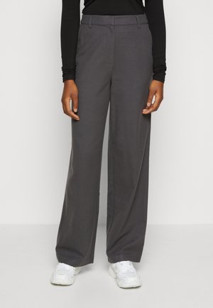 Person trägt lockere, dunkelgraue Hose mit Gürtelschlaufen und weiße Turnschuhe, steht vor einem einfachen hellen Hintergrund.