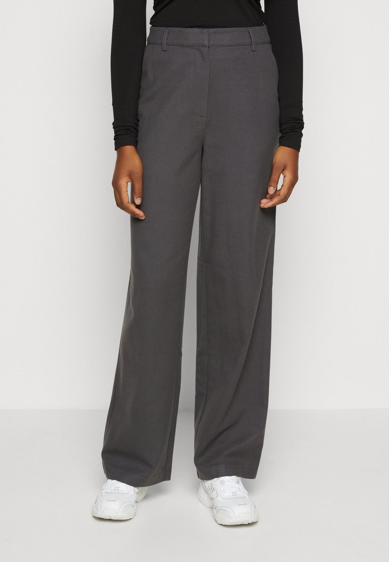 Personne portant un pantalon ample gris foncé avec des passants pour ceinture et des baskets blanches, debout devant un fond clair uni.