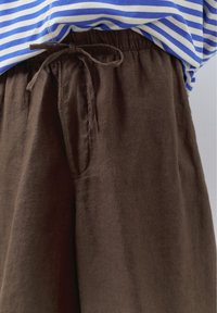 Közeli kép barna lenvászon nadrágról, amelynek a derekán húzózsinór található, kék-fehér csíkos inggel párosítva.