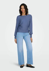 Blauwe geribde trui met brede halslijn, gecombineerd met lichtblauwe jeans met wijde pijpen en zwarte platte schoenen. Effen achtergrond.