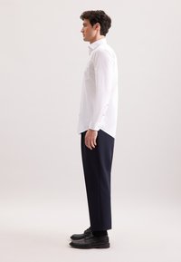 Camicia bianca a maniche lunghe con tasca frontale, polsini con bottoni, abbinata a pantaloni blu navy e scarpe nere, mostrata in una vista laterale.