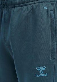 Teal sportbroek van gladde stof. Met een elastische tailleband, zijzakken en een blauwe Hummel-logo print op de bovenbeen.