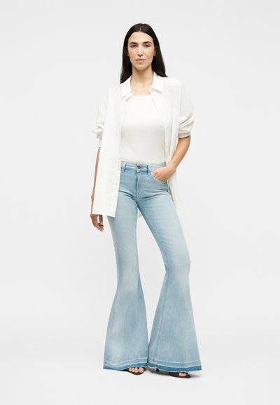 Γυναίκα με μακριά σκούρα μαλλιά που φοράει λευκό πουκάμισο, λευκή φανέλα, ανοιχτό μπλε καμπάνα τζιν και μαύρα σανδάλια με ανοιχτά δάχτυλα, στέκεται με το ένα χέρι στην τσέπη.