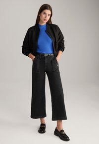 Schwarze Bomberjacke über einem blauen Rippoberteil, kombiniert mit weiten schwarzen Jeans und schwarzen Loafers mit goldfarbenen Hardware-Details.