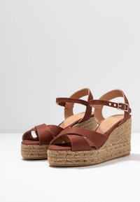Hnědé sandály na platformě se zkříženými pásky, tkanými juty a nastavitelným páskem kolem kotníku se zapínáním na kovovou sponu.