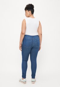 Jeans skinny de color azul oscuro con talle alto, que cuentan con dos bolsillos traseros, combinados con una blusa sin mangas blanca y zapatillas deportivas de color claro.