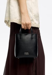 Czarna skórzana torba typu tote o fakturowanej powierzchni, z krótkimi uchwytami i wytłoczonym logo na przodzie. Noszona z jasnym topem.