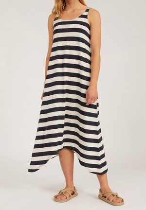 Nainen, joka seisoo päällään mustavalkoinen vaakaraitainen hihaton midi-mekko, jossa on epäsymmetrinen helma, ja beigejä köysisandaaleja.