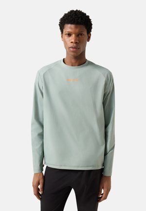 Młody mężczyzna o krótkich, kręconych włosach, ubrany w jasnozieloną koszulę z długim rękawem z pomarańczowym napisem „FIRE + ICE”, stojący na jednolitym tle.