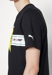 Svart bomull t-shirt med en gul logotyp, vita randiga detaljer och Puma-märke på ärmen. Rund halsringning med korta ärmar.