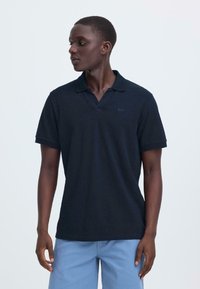 Polo bleu marine foncé avec un col classique, manches courtes et tissu texturé. Marquage subtil sur la poitrine, associé à un short bleu clair.