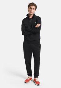 Black zip-up sweatshirt with ribbed cuffs and hem, paired with black jogger pants, accented by logo. Model wears red and white sneakers.