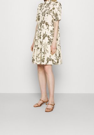 Frau trägt ein locker sitzendes, knielanges cremefarbenes Kleid mit grünem Blumenmuster und kurzen Puffärmeln, kombiniert mit beigefarbenen Sandaletten mit Absatz.