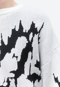 Κοντινό πλάνο λευκού πλεκτού πουλόβερ με ένα έντονο, αφηρημένο μαύρο μοτίβο στο στήθος και τον ώμο.