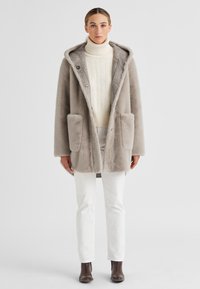 Grauer Kunstpelzmantel mit Kapuze, zwei Vordertaschen und Knopfverschluss, getragen über einem cremefarbenen Rollkragenpullover und weißen Hosen, kombiniert mit braunen Ankle Boots.