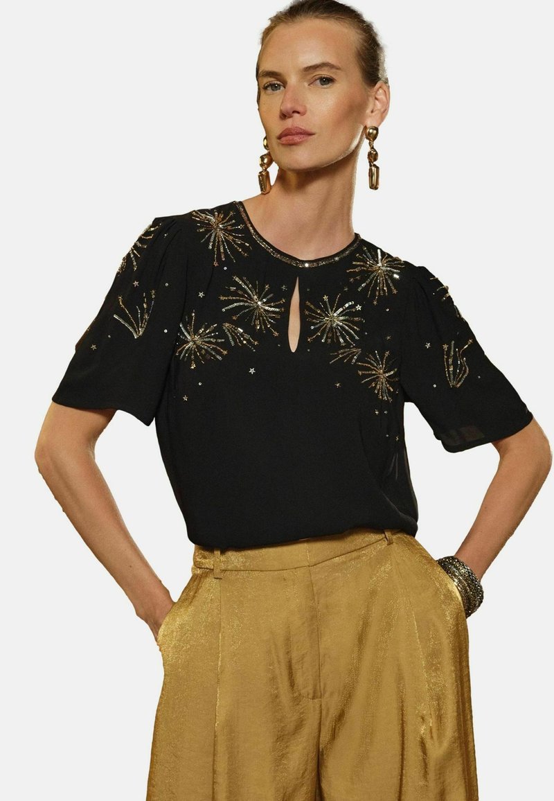 Blusa negra con mangas cortas abullonadas, que presenta bordado dorado y abalorios en un patrón de explosión de estrellas; el escote tipo ojo de cerradura añade detalle. Pantalones dorados de cintura alta visibles.