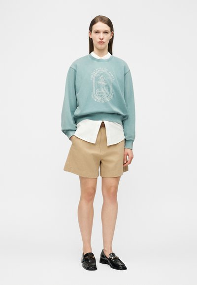 Γυναίκα που στέκεται φορώντας πετρόλ φούτερ με σχέδιο πάνω από λευκό πουκάμισο, μπεζ σορτσάκια μήκους έως τα γόνατα και μαύρα μοκασίνια σε απλό γκρι φόντο.