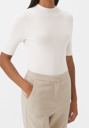 Frau trägt ein eng anliegendes weißes kurzärmeliges Oberteil und hoch taillierte beige Hose, mit einer Hand in der Tasche vor einfachem Hintergrund.