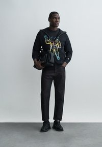 Férfi áll, fekete cipzáras kapucnis pulóvert visel, amelyen színes űrhajós grafika található, sötét nadrágot és fekete sportcipőt hord, egyik kezében fekete táskát tart.