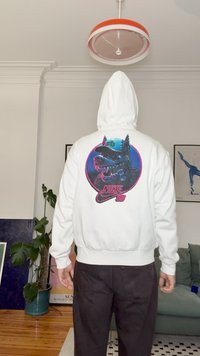Vit hoodie med en stor grafik av en varg som bär keps på ryggen. Har elastiska muddar och midjeresår; minimalistisk design.