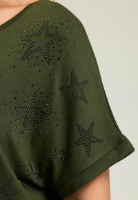 Top de manga corta color verde oliva con un corte relajado, que presenta patrones de estrellas negras contorneados con brillantes strass. Textura de tela suave.