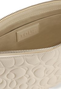 Bolso de color crema con patrones circulares en relieve, interior forrado y cierre con cremallera. Presenta una etiqueta en relieve "TOUS" en su interior.