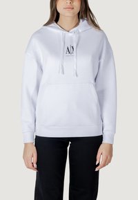 Sudadera blanca de tejido suave, con un bolsillo canguro frontal, capucha con cordón y un logo en el pecho. Combinada con pantalones negros.