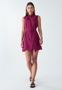 Fuksijos spalvos be rankovių suknelė su apykakle, priekyje su mygtukais ir juosmens raiščiu. Apatinėje dalyje yra horizontalios juostelės, o siluetas - laisvas.