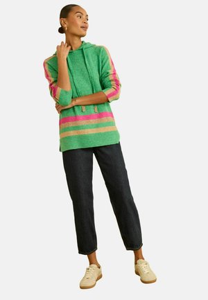 Grüner Kapuzenpullover mit mehrfarbigen Streifen in Pink und Gold. Kombiniert mit dunkelblauer Denim-Hose und hellen Sneakers.